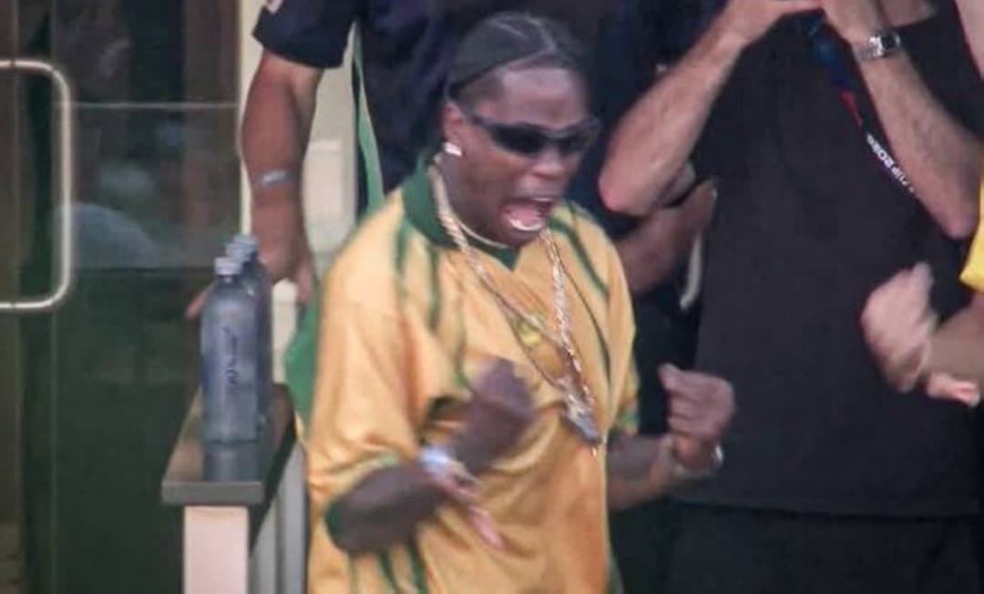Travis Scott wearing a yellow and green Brazil shirt celebrates in the stands of MetLife Stadium during the 2026 World Cup match between Morocco and Brazil.