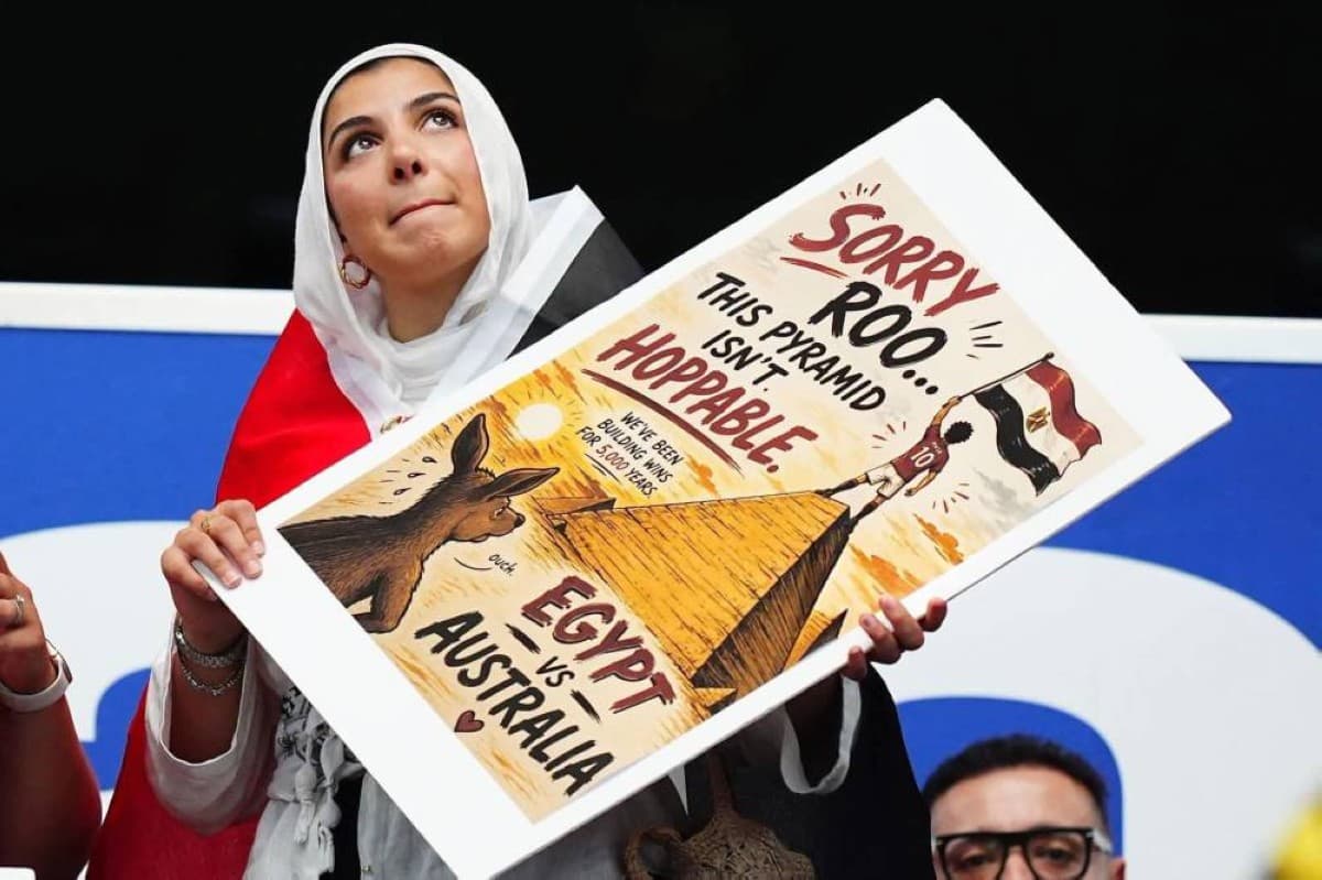 An Egyptian female fan wearing a hijab and the Egyptian flag holds up a hand-painted banner at the 2026 FIFA World Cup, depicting an Egyptian player raising his country's flag atop a pyramid while a kangaroo looks on.
