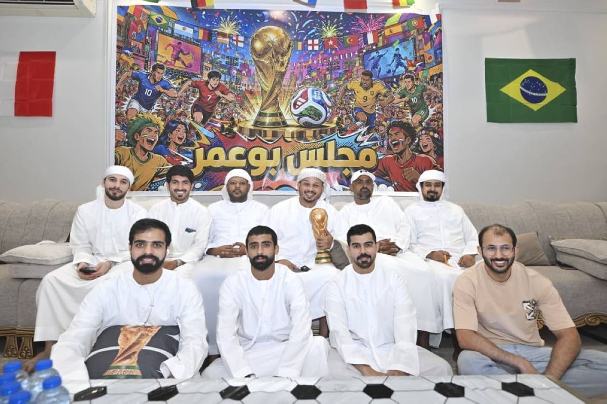 A decorated home majlis in Abu Dhabi filled with national team flags hanging from the ceiling and walls, football jerseys, and a large screen, set up by Emirati fan Mohamed Al Mashjari for watching World Cup 2026 matches with friends.