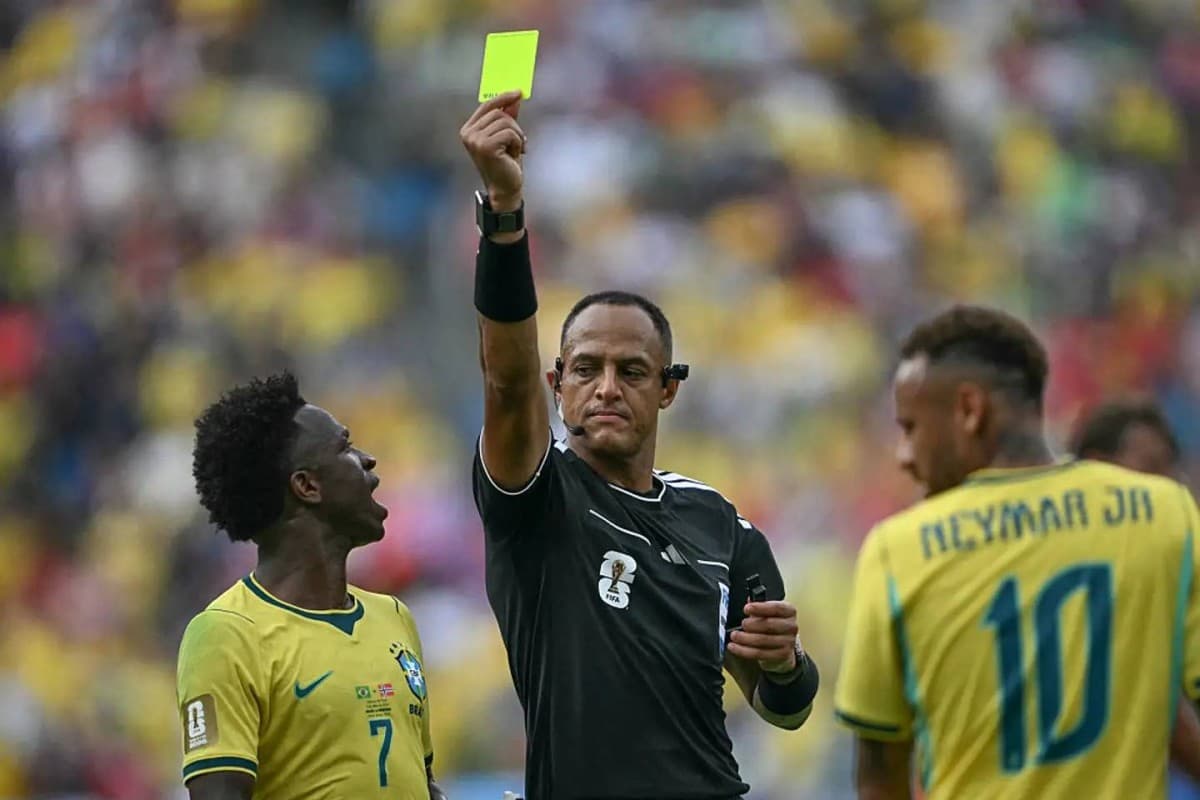 A referee holds up a yellow card during a World Cup 2026 match, with players from two national teams visible in the background.