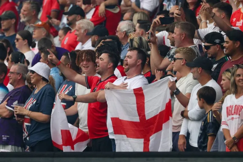 England fans in the stands at a World Cup match, with some holding flags and scarves in the team's colours.