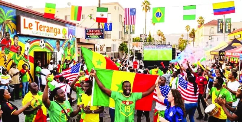 Crowds of football fans gathered in a vibrant immigrant neighbourhood street in a major American city, watching a World Cup match on a large outdoor screen.